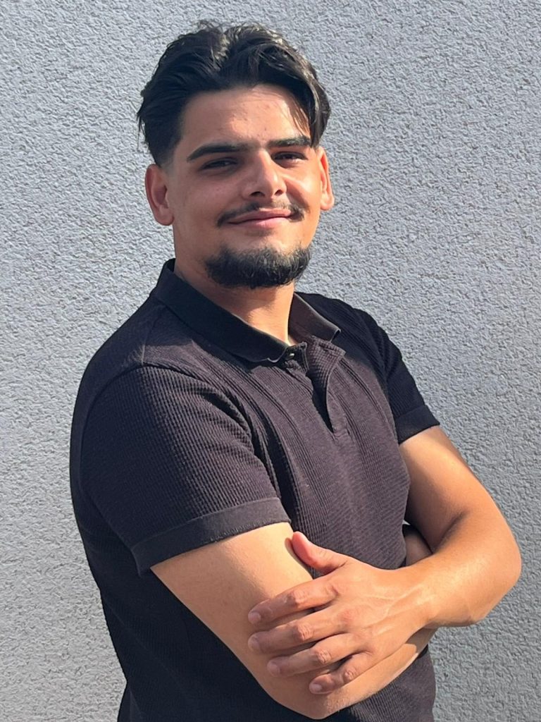 Clean Police Luan Shabani Junger Mann mit kurzem Haar in schwarzem Polohemd, lächelt und steht mit verschränkten Armen.